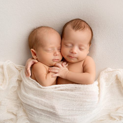 Tiny newborn twins boys in white cocoons on a white background. A newborn twin sleeps next to his brother. Newborn two twins boys hugging each other. Professional studio photography.
